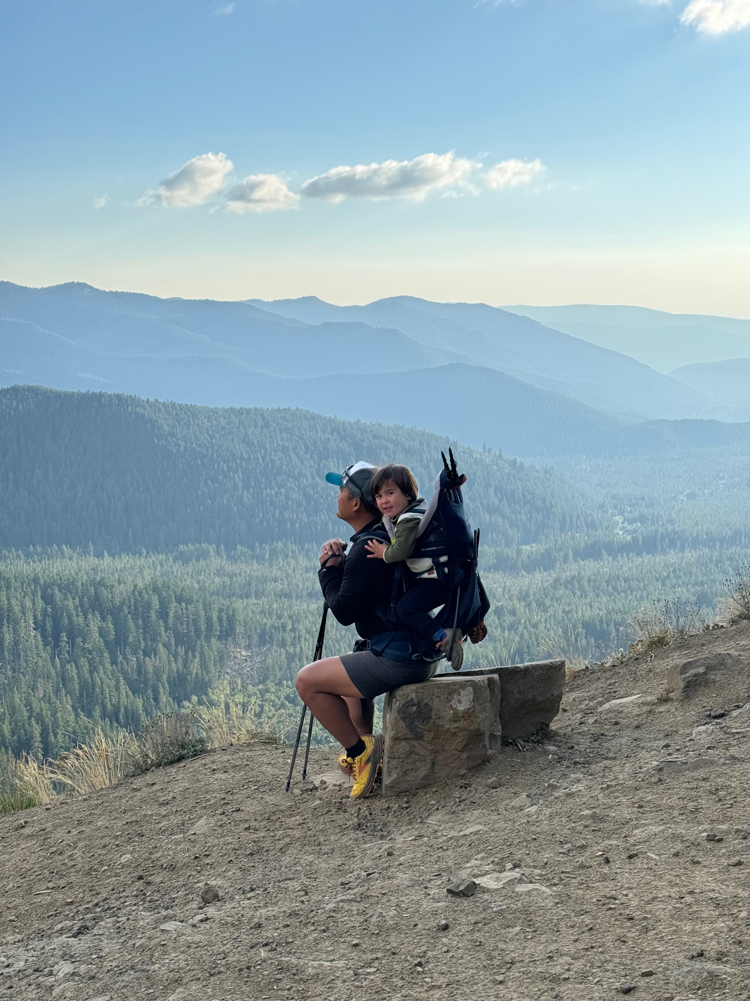 Adult and child sitting on a log on a hiking trail, looking left to a view of mountains in the distance