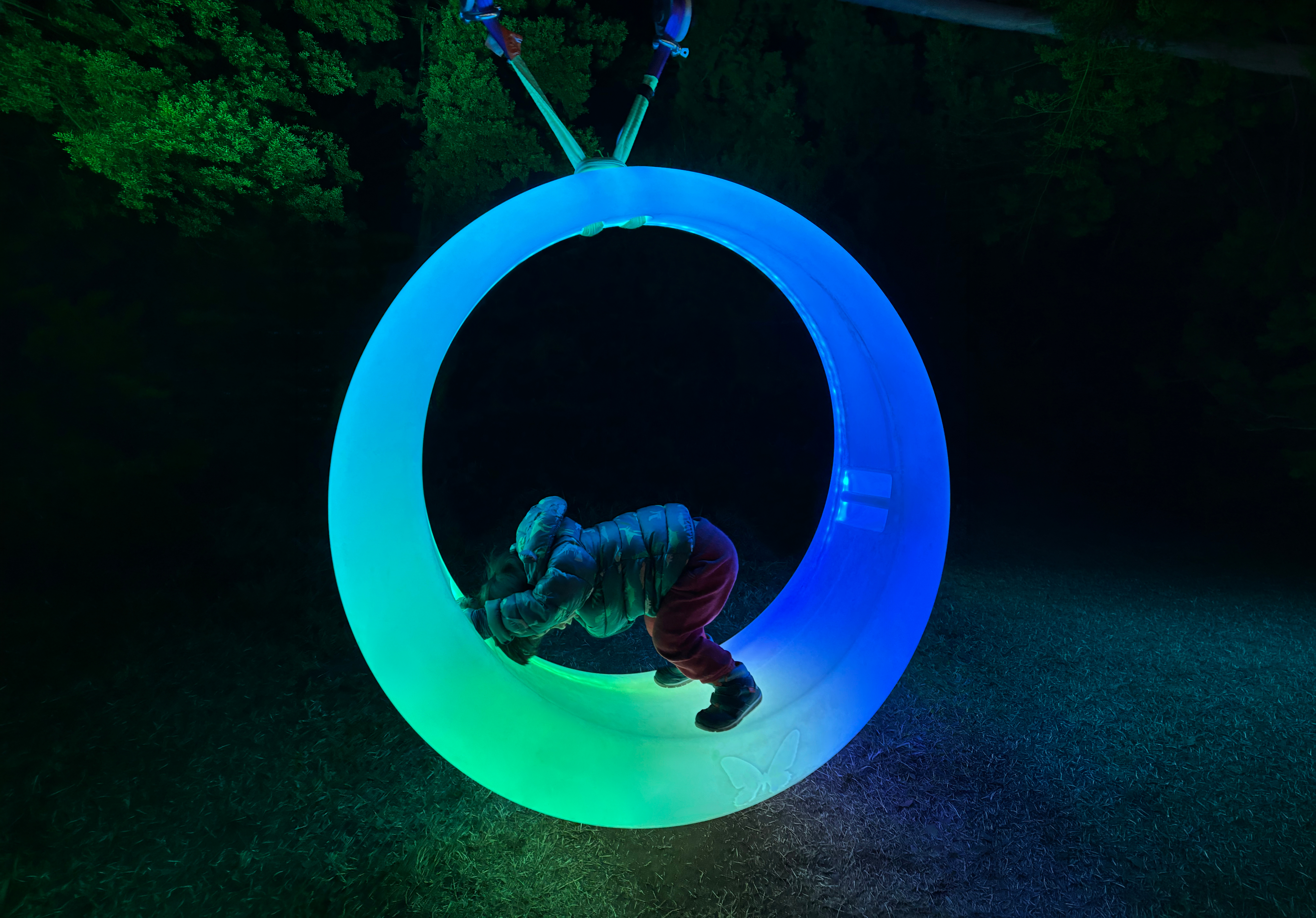 A child inside a suspended plastic ring swing, lit in a gradient of green and purple